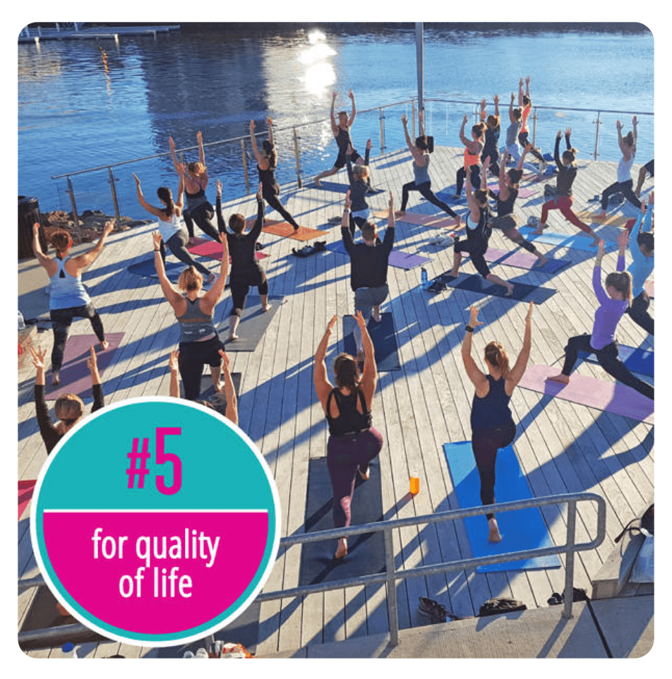 An outdoor yoga class in Connecticut. Participants stand on their yoga mats on a sunny deck next to a river.
